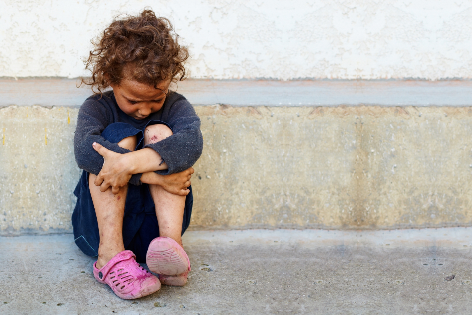 poor, sad little child girl sitting against the concrete wall