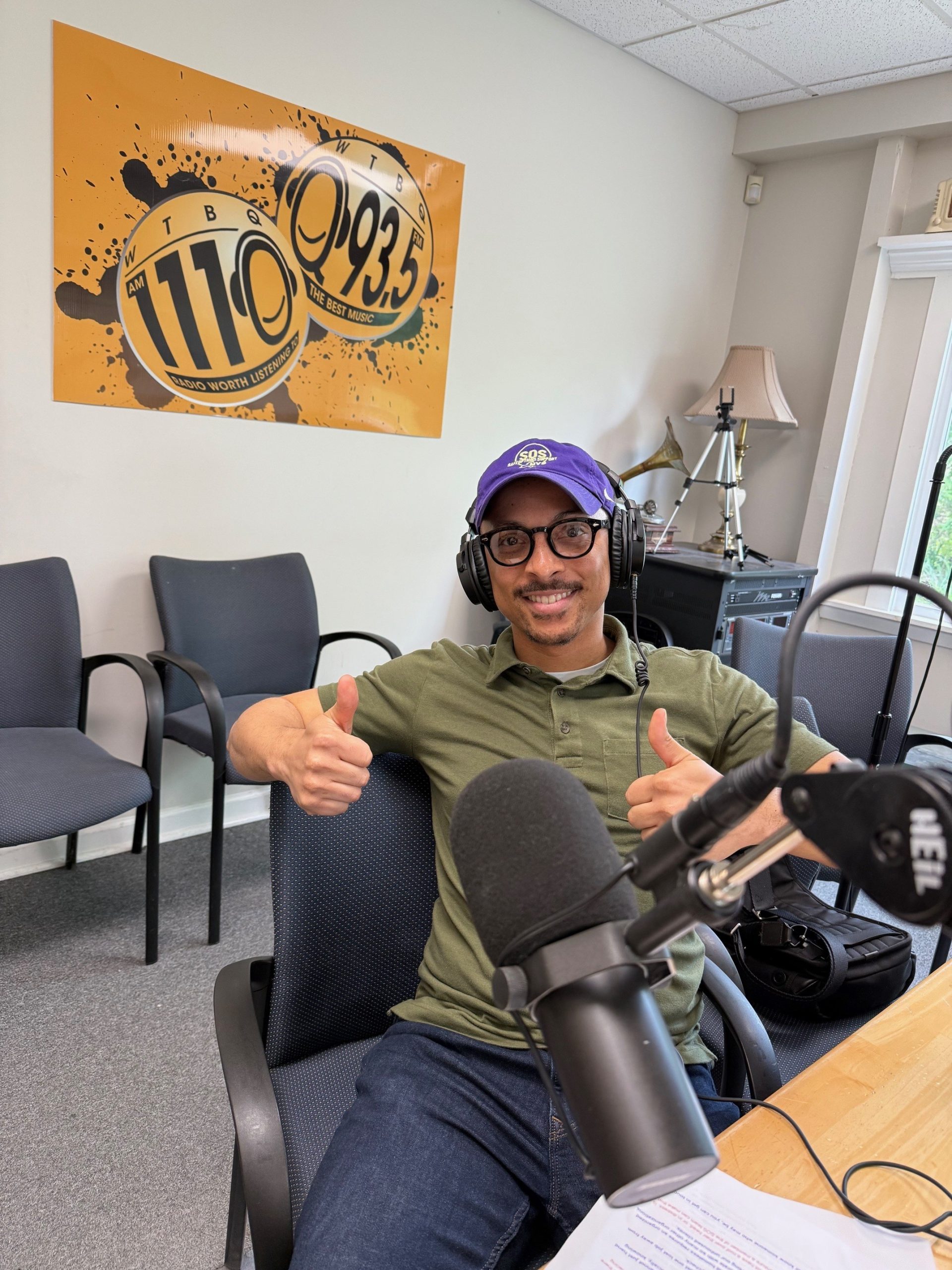 A man wearing a purple SOS hat, glasses, and headphones sits in a radio studio, smiling and giving two thumbs up. He is seated in front of a large microphone and a desk with printed notes. Behind him is a sign for WTGB 1110 AM / 93.5 FM and a few empty chairs.