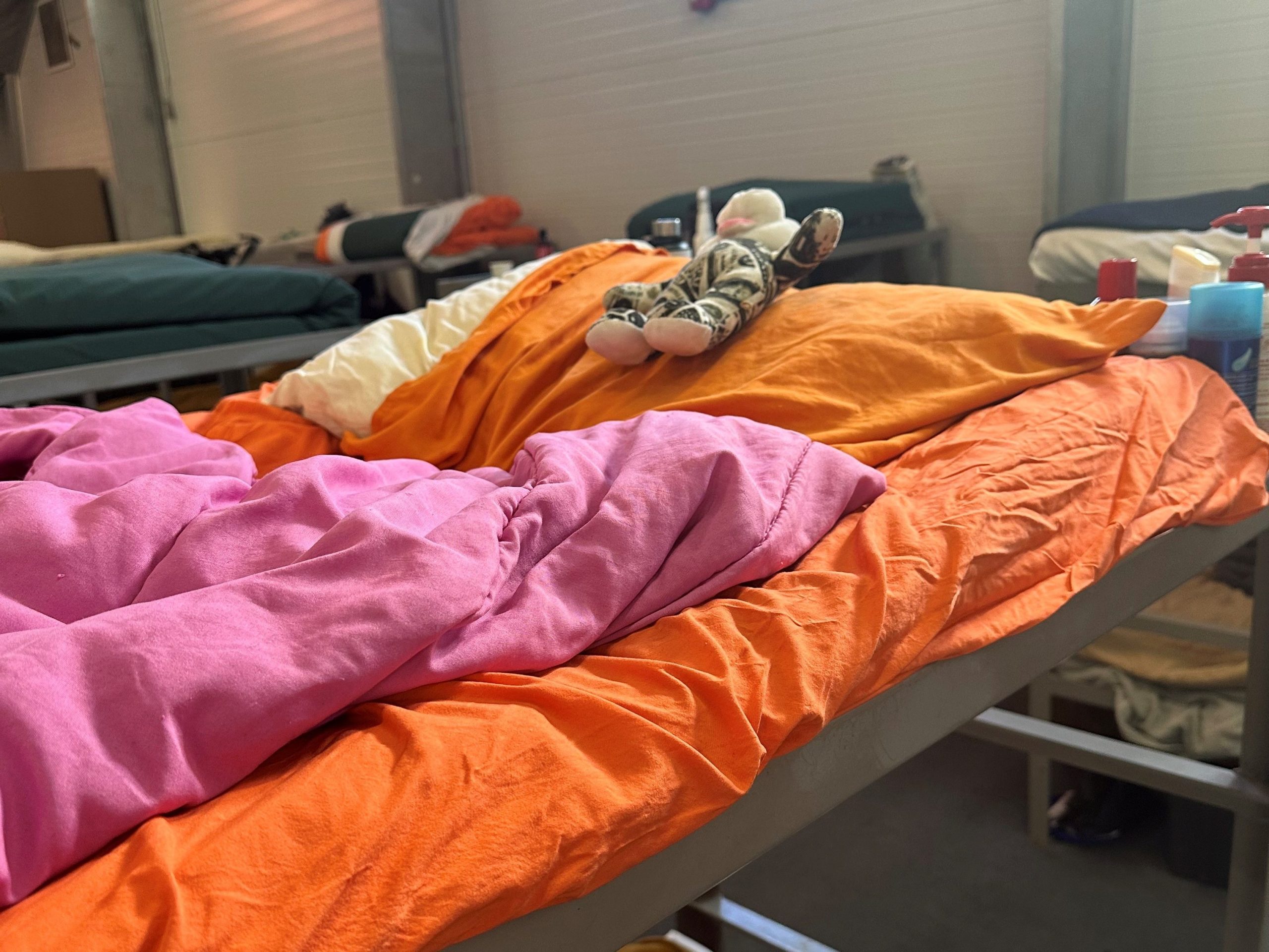A metal bunk bed in a shelter setting, made up with an orange pillow, pink comforter, and a small stuffed animal resting on top.