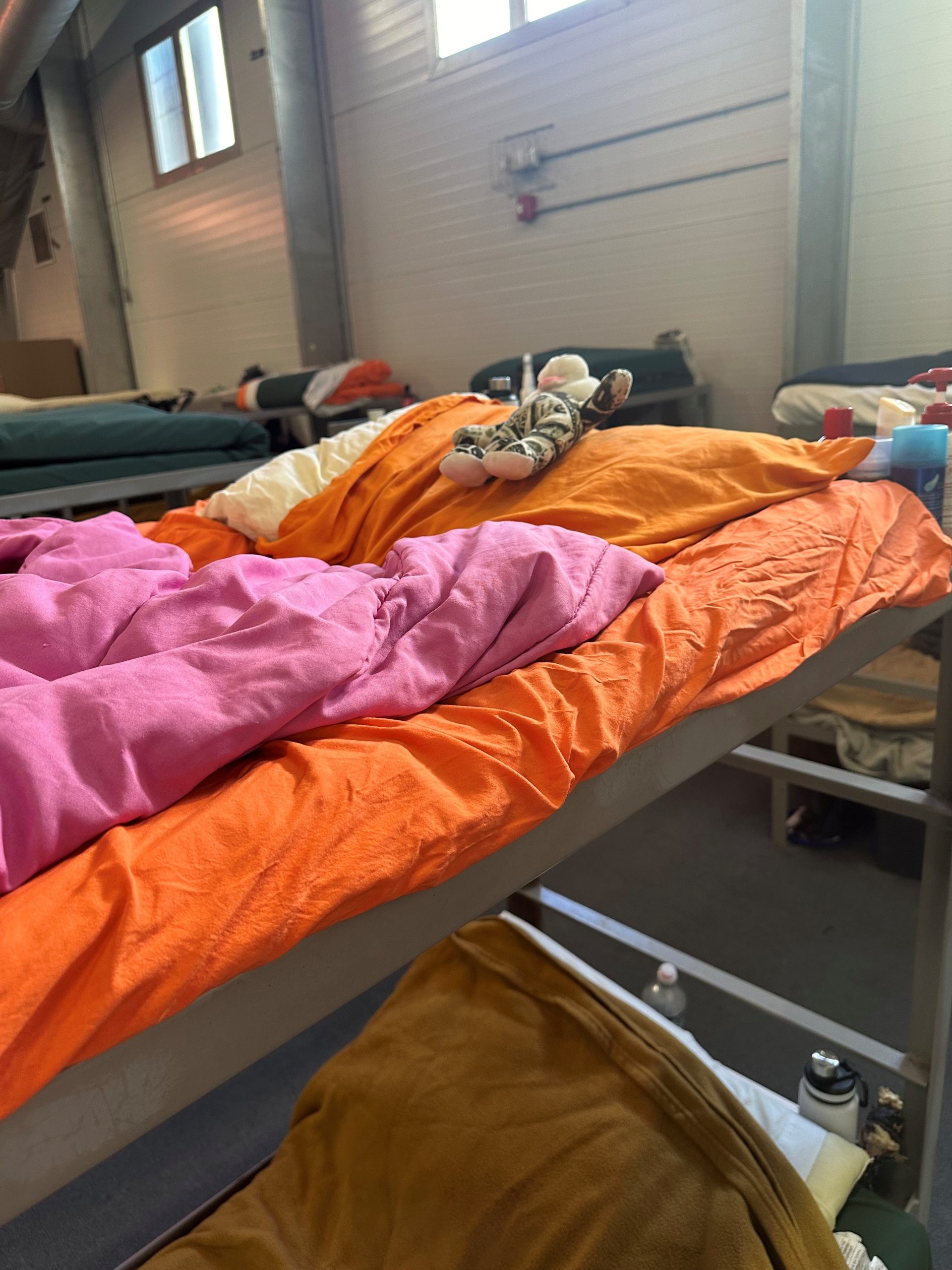 A metal bunk bed in a shelter setting, made up with an orange pillow, pink comforter, and a small stuffed animal resting on top.