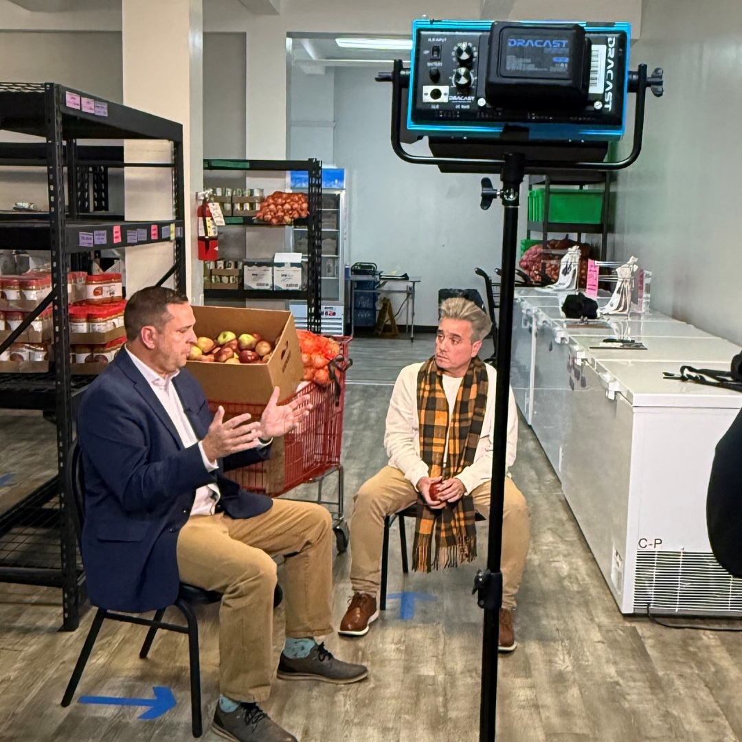 Two people sit in a food-pantry room having an on-camera discussion. One speaks with hands raised while the other listens. Shelves of produce, a box of apples, and freezers line the background, with a studio light in the foreground.